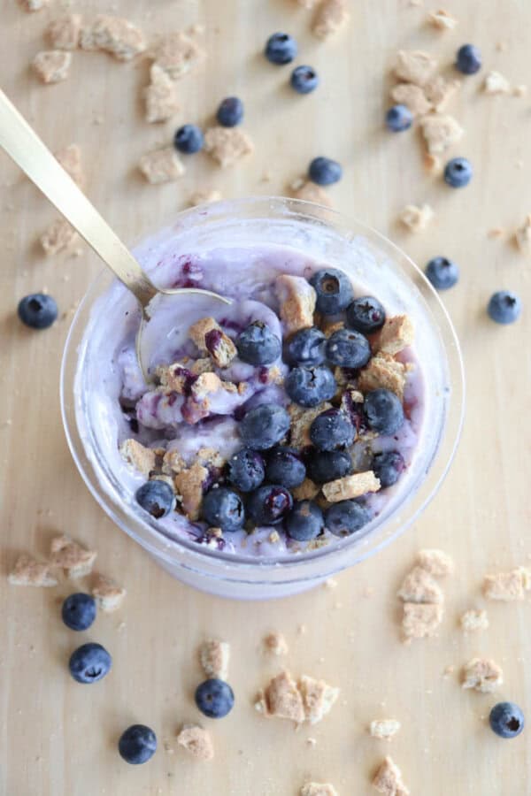 A bowl of blueberry ice cream topped with fresh blueberries and crumbled biscuit pieces evokes the creamy delight of blueberry cheesecake ice cream. A gold spoon rests inside, while extra blueberries and biscuit crumbs are scattered on the wooden surface around the bowl.