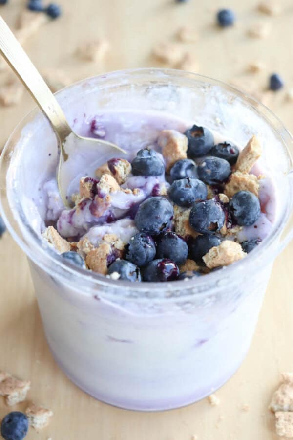 A pint filled with ice cream mixed with blueberries and crumbled granola, reminiscent of blueberry cheesecake ice cream. The yogurt has a swirl of purple, indicating the presence of blueberries. A spoon rests inside, while a few berries and bits of granola are scattered around.
