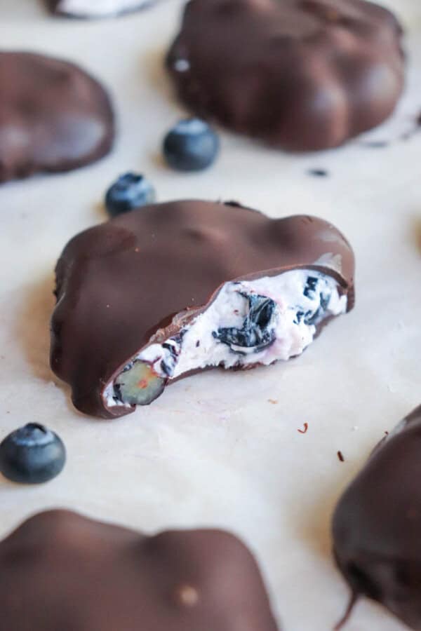 Chocolate-covered blueberry treats filled with cream, one split open to show blueberries inside. Other whole treats and scattered blueberries surround it on a parchment-lined surface.