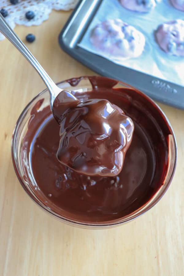 A spoonful of rich, melted chocolate is held above a glass bowl filled with more chocolate. In the background, a tray with cookies partially coated in chocolate is visible on a wooden surface.