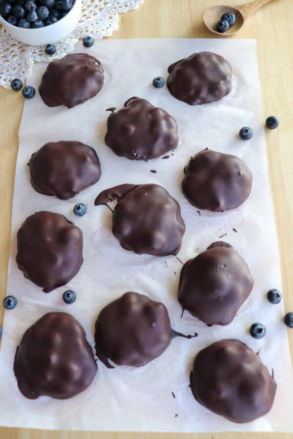Ten chocolate-covered clusters are arranged on a sheet of parchment paper with scattered blueberries around. A small bowl of blueberries and a spoon are visible in the background on a wooden surface.