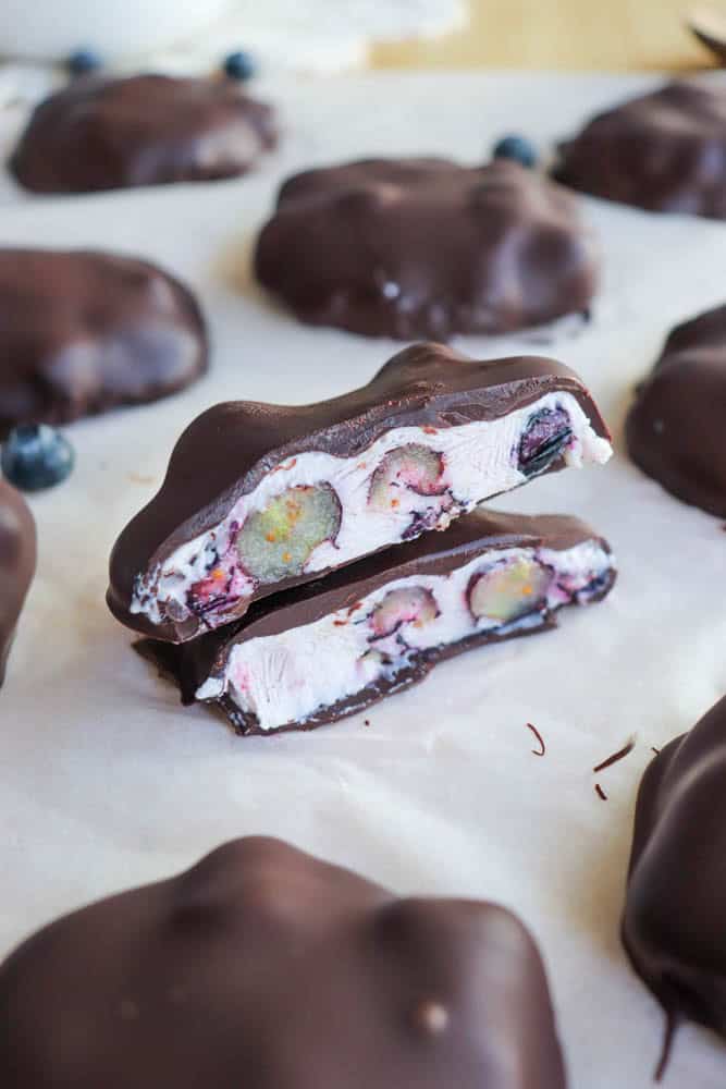 Chocolate-covered treats with a creamy filling and blueberries are displayed on parchment paper. One treat is halved, revealing the inside with white cream and blueberries. Background shows more treats and blurred blueberries.