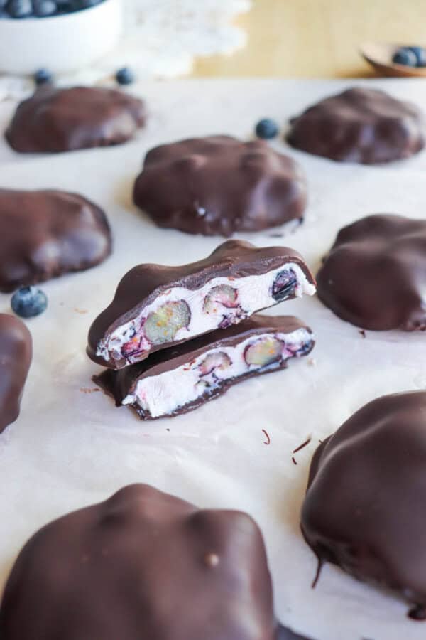 Chocolate-covered treats are arranged on parchment paper. One is cut in half, revealing a creamy filling with whole blueberries inside. A small bowl with more blueberries is in the background.