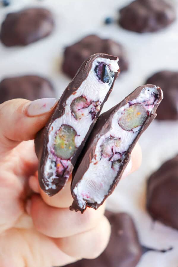 Close-up of a hand holding a halved chocolate-covered treat revealing white marshmallow filling and whole blueberries inside. Other similar treats are blurred in the background.