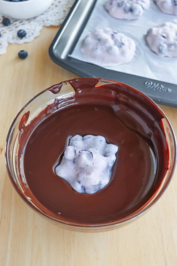 A bowl of melted chocolate with a blueberry-filled, white mixture partially submerged. In the background, a baking tray with more similar white and blueberry mixtures on a wooden surface.