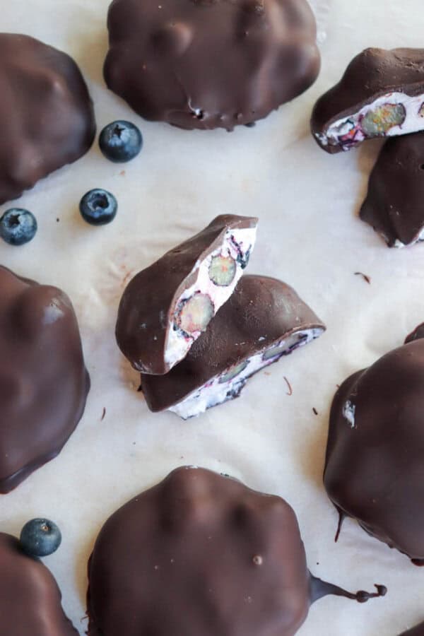 Chocolate-covered treats with a cut piece showing a creamy filling and blueberries inside. Several whole blueberries are scattered around on a parchment surface.