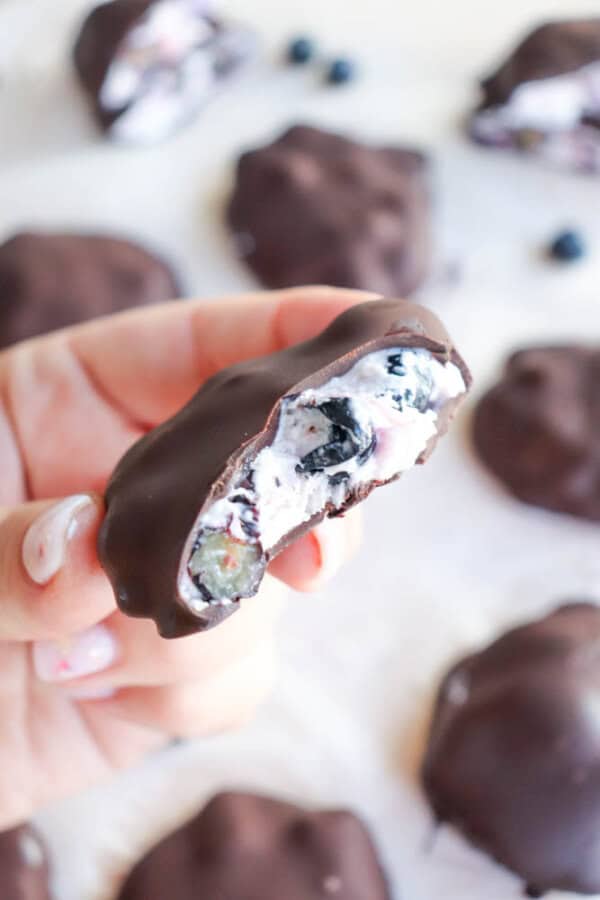 A hand holding a chocolate-coated treat with a bite taken out, revealing a creamy filling and blueberries inside. More chocolate-coated treats are blurred in the background.