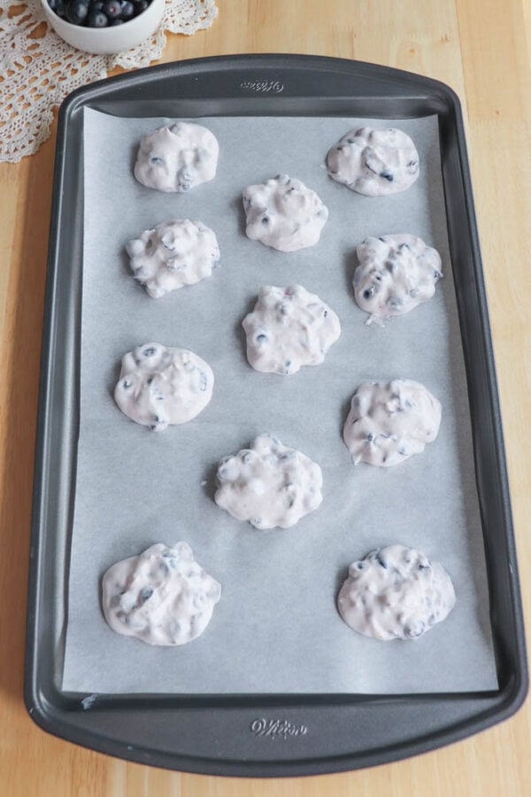 A baking tray lined with parchment paper holds twelve dollops of cookie dough mixed with blueberries, ready to be baked. A small bowl of blueberries is in the background on a wooden surface.