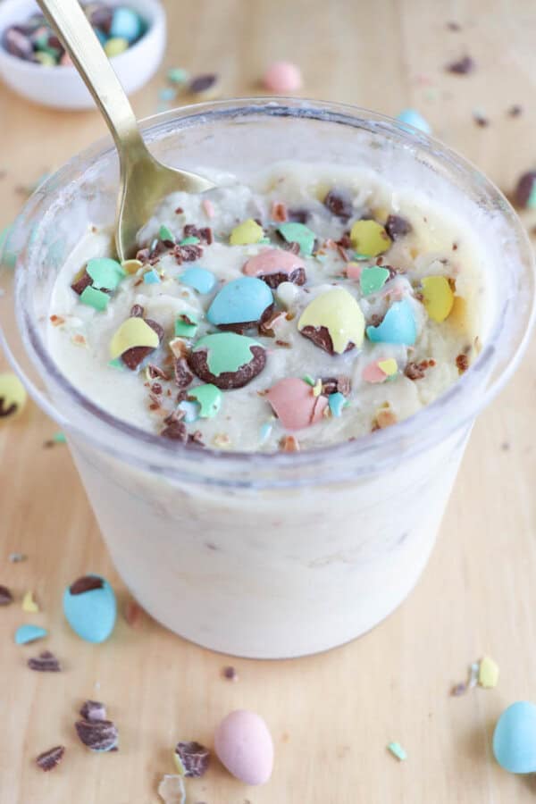 A cup of creamy dessert reminiscent of bunny tracks ice cream is topped with colorful mini chocolate eggs and chocolate shavings. A spoon is dipped into the treat, while more candy eggs are scattered on the wooden table surface.