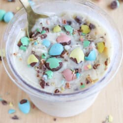 A dessert cup filled with creamy white ice cream ice cream, topped with crushed pastel-colored chocolate candies. A spoon is placed in the cup, and more candy pieces are scattered on the wooden surface around the cup.