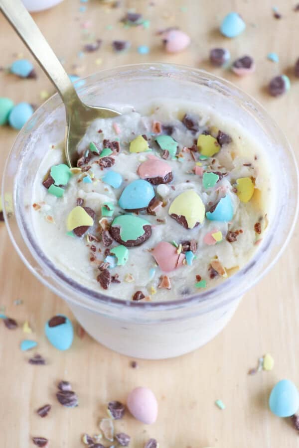 A dessert cup filled with creamy white ice cream ice cream, topped with crushed pastel-colored chocolate candies. A spoon is placed in the cup, and more candy pieces are scattered on the wooden surface around the cup.