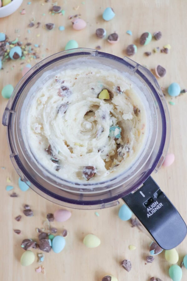 A food processor filled with a creamy mix of bunny tracks ice cream and colorful candy pieces. Surrounding the processor on the wooden surface are scattered pieces of these vibrant candies in pastel blue, pink, yellow, and brown.