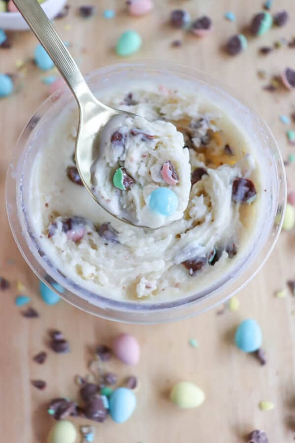 A spoon dips into a creamy Bunny Tracks ice cream mixture with chocolate chips and colorful candy pieces in a container. More candy and chocolate chips are scattered on the wooden surface around the container.