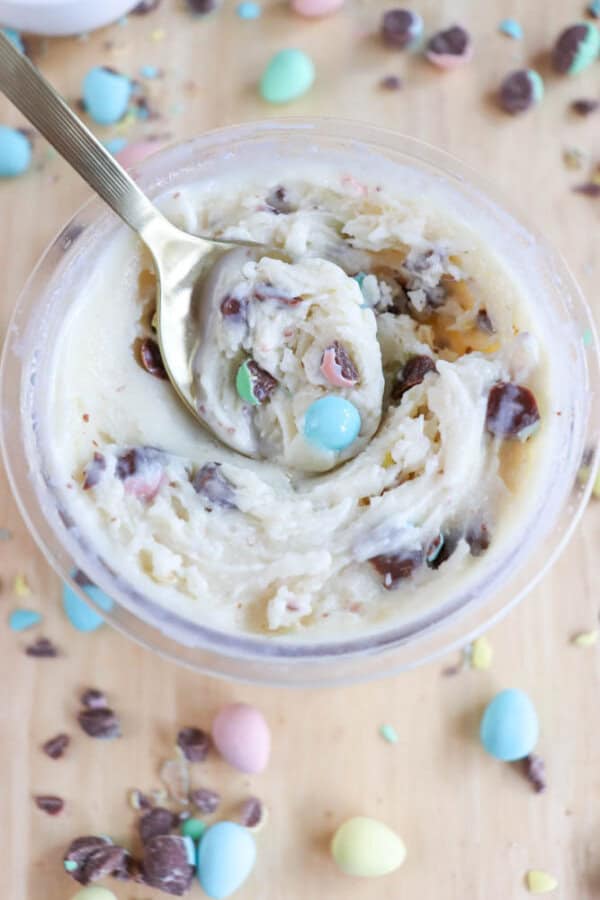 A spoon rests in a jar of cookie dough, reminiscent of bunny tracks ice cream. The dough is dotted with chocolate chips and colorful candy-coated chocolate eggs. More candies and chocolate chips are scattered on the light wooden surface around the jar.