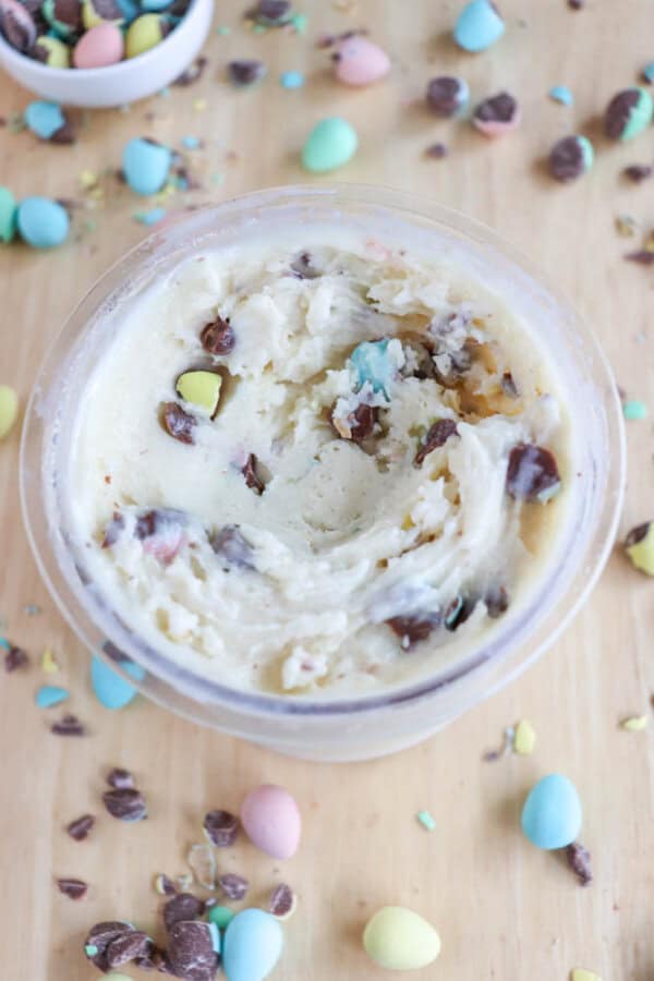 A jar of cookie dough is topped with colorful candy-coated chocolate pieces, reminiscent of bunny tracks ice cream, in pastel shades of blue, pink, and yellow. Surrounding the jar, more scattered candies create a festive look on the wooden surface.