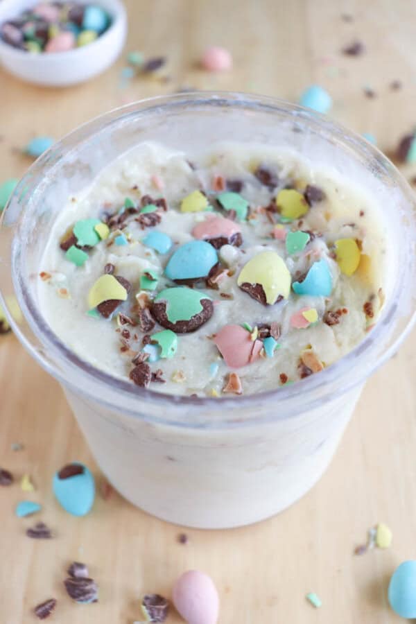 A small container filled with creamy bunny tracks ice cream, topped with colorful, crushed candy pieces on a light wooden surface. More candy pieces are scattered around, and a small bowl with candies is visible in the background.