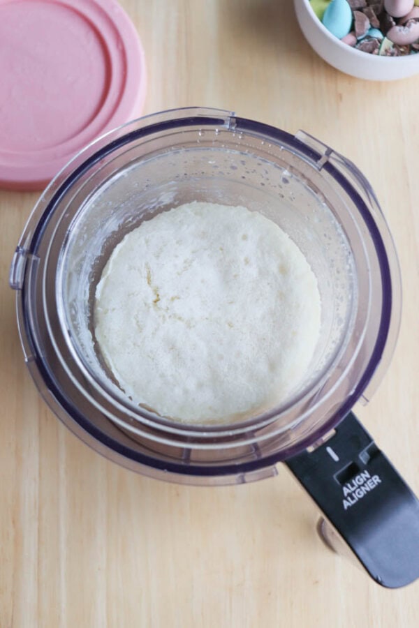 A top view of a food processor containing a smooth, white dough on a wooden surface, reminiscent of the creamy base of bunny tracks ice cream. A bowl filled with colorful candies is partially visible in the background, adding vibrant contrast to the serene scene.