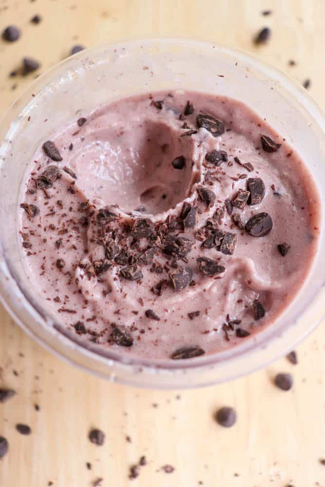 A close-up of a creamy, thick pink smoothie in a bowl topped with dark chocolate chunks. Some chocolate pieces are scattered around the bowl on a light wooden surface.