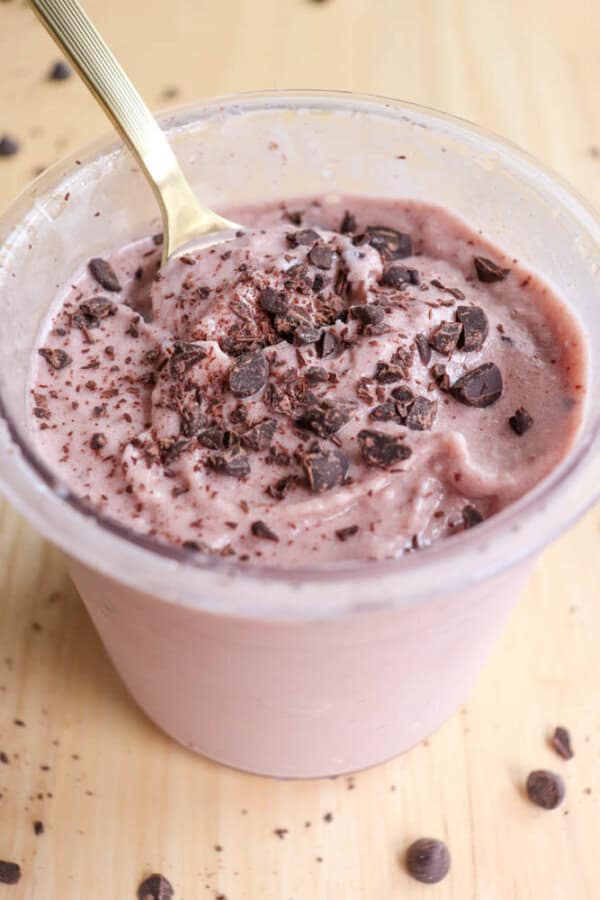 A plastic cup filled with a creamy, pink dessert topped with chocolate chunks, placed on a wooden surface. A golden spoon is inserted into the dessert, and a few chocolate pieces are scattered around the cup.