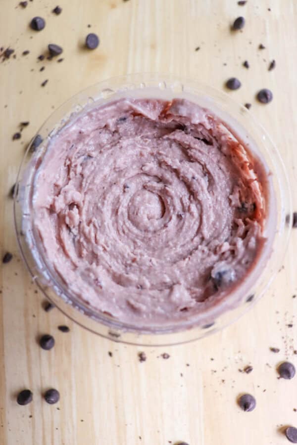 A bowl of chocolate chip cookie dough is placed on a wooden surface. Chocolate chips are scattered around the bowl, adding to the desserts appeal. The dough has a smooth, creamy texture with visible chocolate chips embedded in it.