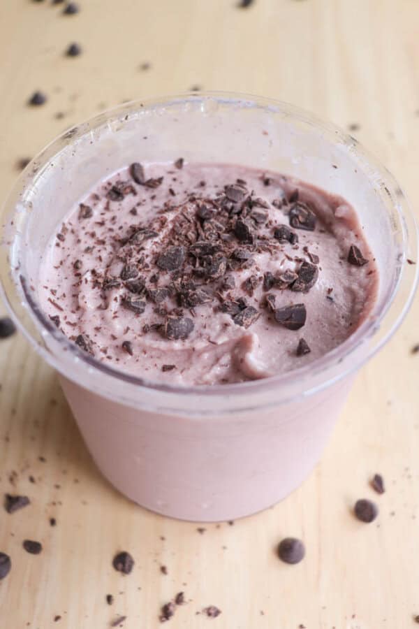 A clear plastic cup filled with creamy chocolate ice cream topped with chocolate shavings. Scattered chocolate chips surround the cup on a light wooden surface.