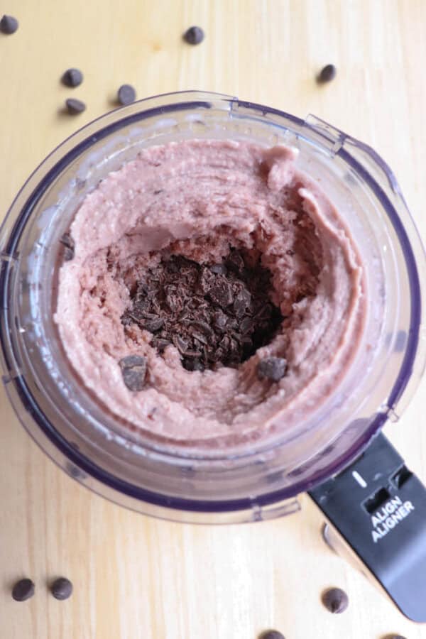 A food processor containing a creamy, pink mixture with added chocolate chips and chopped chocolate. The appliance is sitting on a wooden surface.