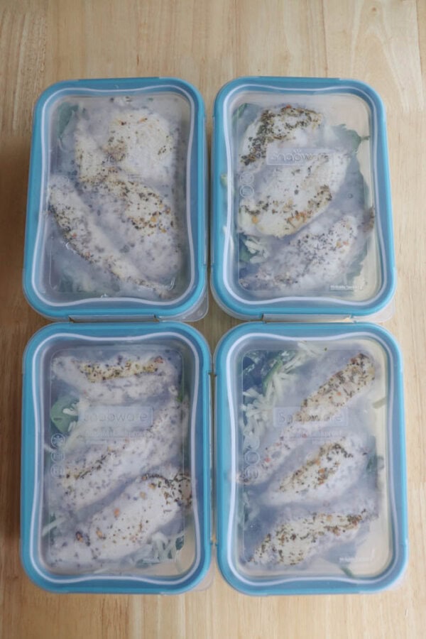 Four glass meal prep containers with blue lids, filled with grilled chicken slices and a hint of spinach pesto bake, are neatly arranged on a wooden surface.
