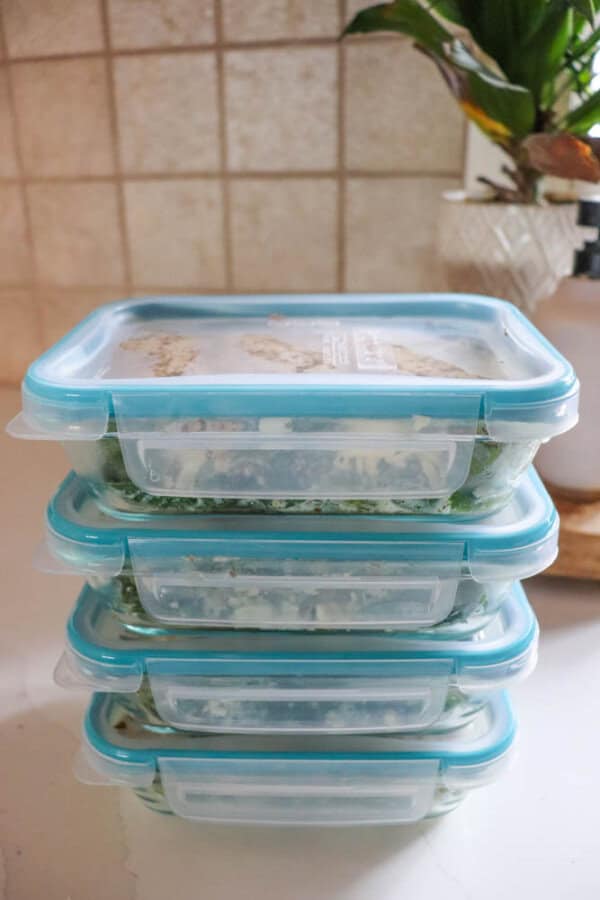 Four stacked glass meal prep containers with blue lids rest on a white counter, each brimming with a vibrant chicken spinach pesto bake. A plant in a white pot adds a touch of life in the background against the tiled backsplash.