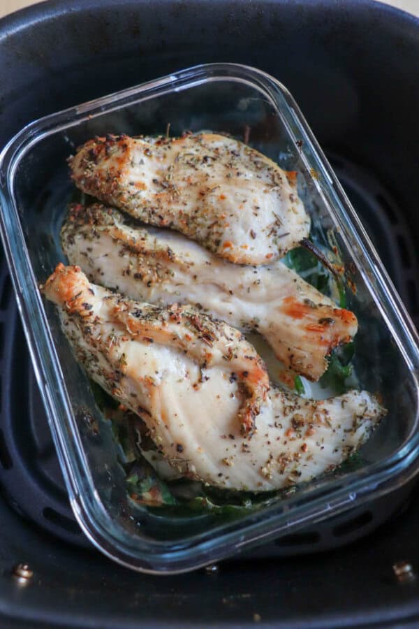 A glass container cradles three pieces of seasoned, cooked chicken breast in an air fryer. This delectable chicken spinach pesto bake is topped with vibrant herbs and spices, showcasing a golden, textured appearance thats simply irresistible.