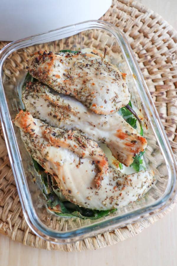 Grilled chicken breast slices seasoned with herbs rest elegantly in a glass container on a woven mat. The addition of leafy greens and hints of pesto creates a fresh, appetizing presentation.