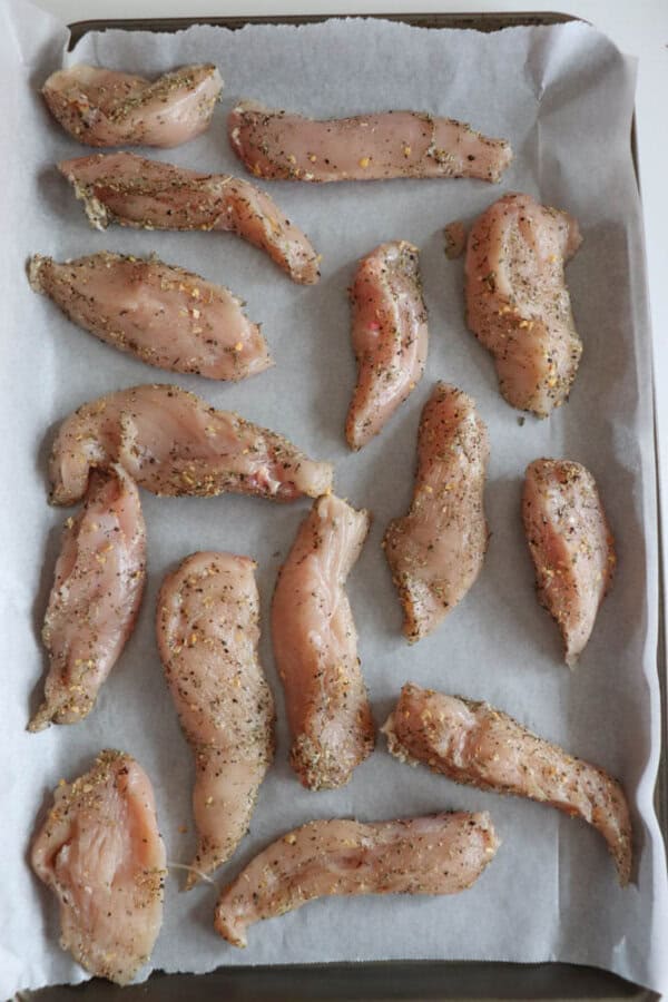 Raw, seasoned chicken strips are placed on a parchment-lined baking tray, ready to be transformed into a delicious chicken pesto bake. The seasoning, along with the vibrant green spinach pesto, is evenly distributed across the pieces for a flavorful dish.