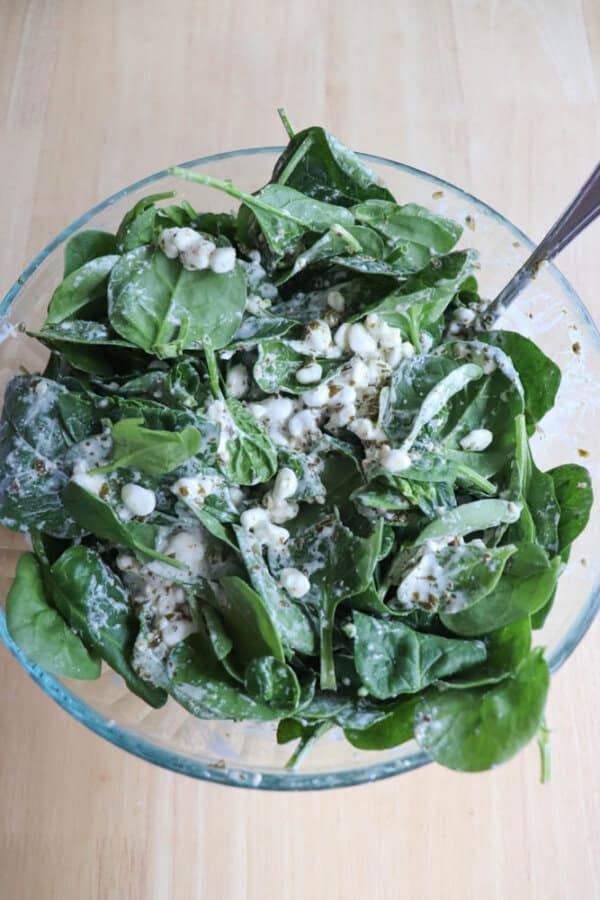 A clear glass bowl brimming with fresh green spinach leaves, crumbled blue cheese, and creamy dressing sits on a light wooden surface. Complementing the enticing mix, a metal spoon awaits to serve up the chicken spinach pesto bake-inspired flavors within.