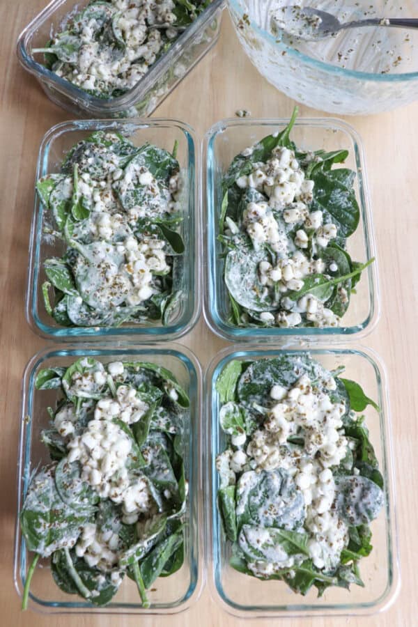 Four glass containers are filled with fresh spinach leaves, crumbled feta cheese, and a creamy dressing, reminiscent of a chicken spinach pesto bake. A mixing bowl with a spoon, containing remnants of the dressing, is partially visible in the corner.