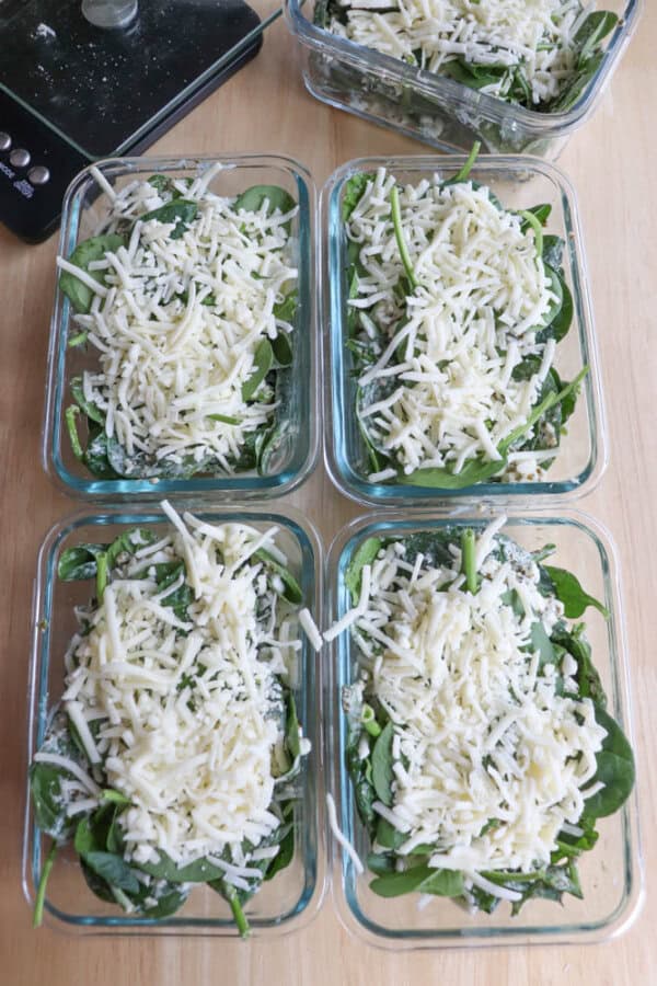 Five glass containers filled with fresh spinach leaves and shredded cheese are arranged on a wooden surface, whispering of the chicken spinach pesto bake soon to come. A black kitchen scale peeks from the top left corner, poised for precision.