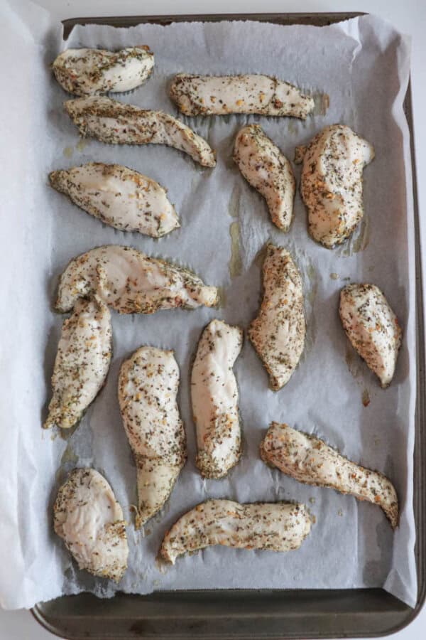Baked chicken tenderloins seasoned with herbs and spices are arranged on a parchment-lined baking sheet. The tenderloins, part of a delightful chicken spinach pesto bake, boast a lightly golden appearance.