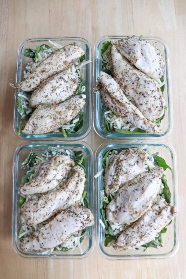 Four glass containers filled with seasoned grilled chicken breasts on a bed of mixed greens and drizzled with a touch of spinach pesto, placed on a wooden surface.