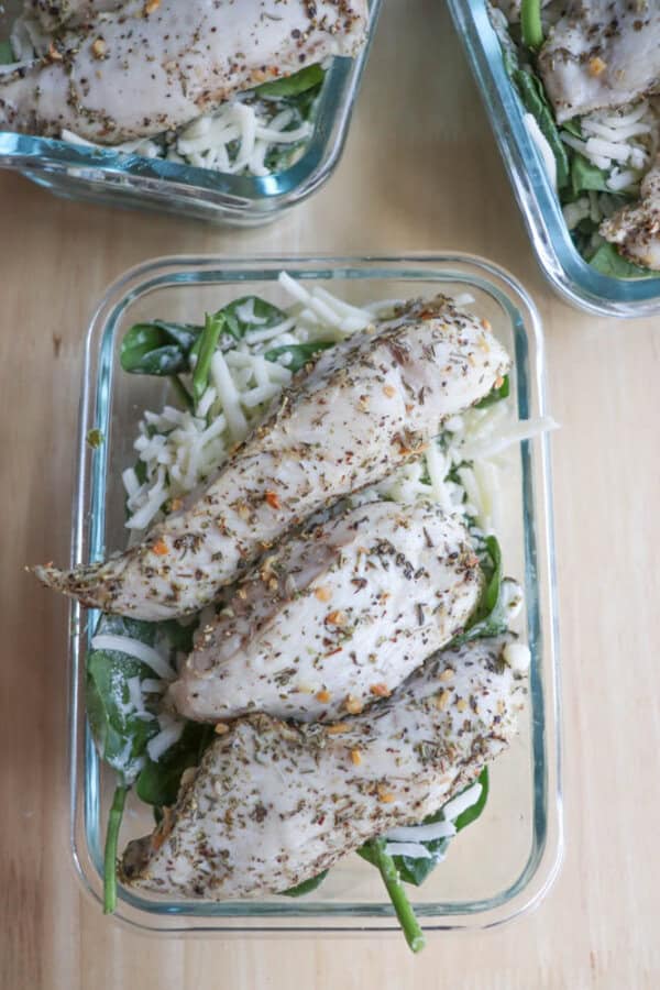 Glass containers filled with seasoned chicken breast fillets rest on a bed of spinach and grated cheese, infused with a hint of pesto. The dish is beautifully displayed on a wooden surface.