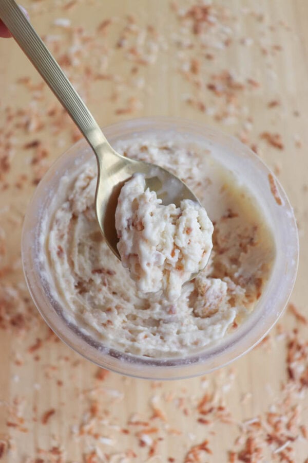 A spoon scooping coconut cream pie ice cream from a plastic container. The mixture has a textured appearance with visible bits, possibly coconut. The container is placed on a wooden surface sprinkled with similar bits.