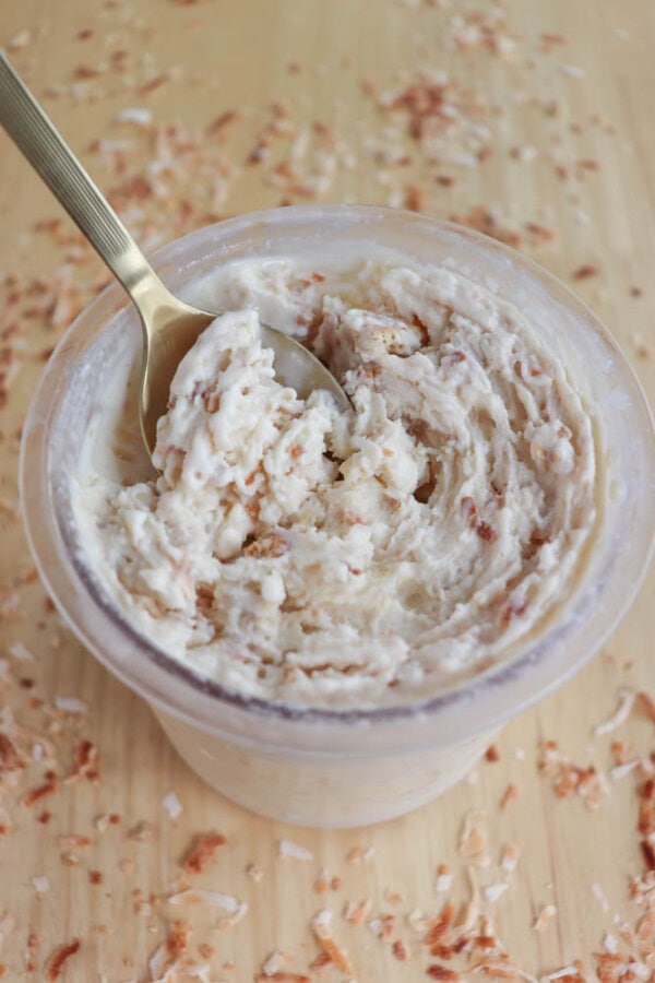 A cup filled with creamy coconut cream pie ice cream topped with shredded coconut, placed on a wooden surface scattered with additional coconut flakes. A gold spoon is dipping into the dessert.