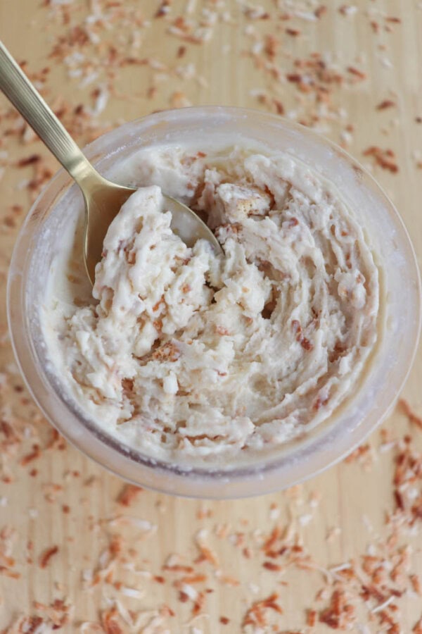 A clear bowl of creamy coconut-flavored butter, featuring visible coconut shavings, with a gold spoon scooping from the top. The wooden surface underneath is scattered with additional coconut flakes.