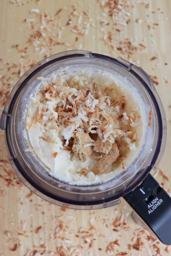 Top view of a food processor containing a creamy mixture topped with crumbled graham cracker pieces and toasted coconut flakes, against a wooden background with scattered coconut flakes.