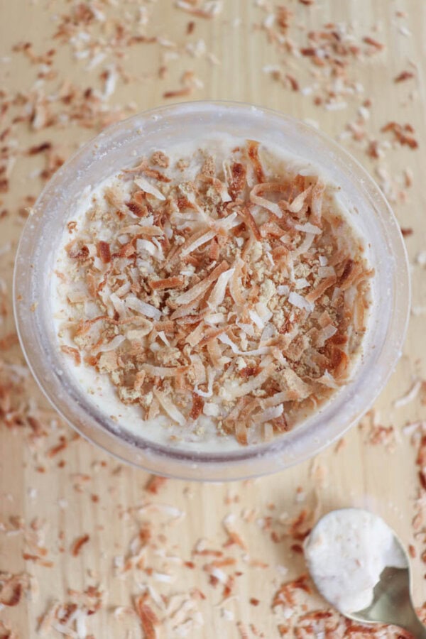 A glass of creamy coconut cream pie ice cream topped with toasted coconut flakes and crumbled graham crackers, placed on a wooden surface scattered with more coconut flakes. A spoon with a bit of dessert rests beside the glass.