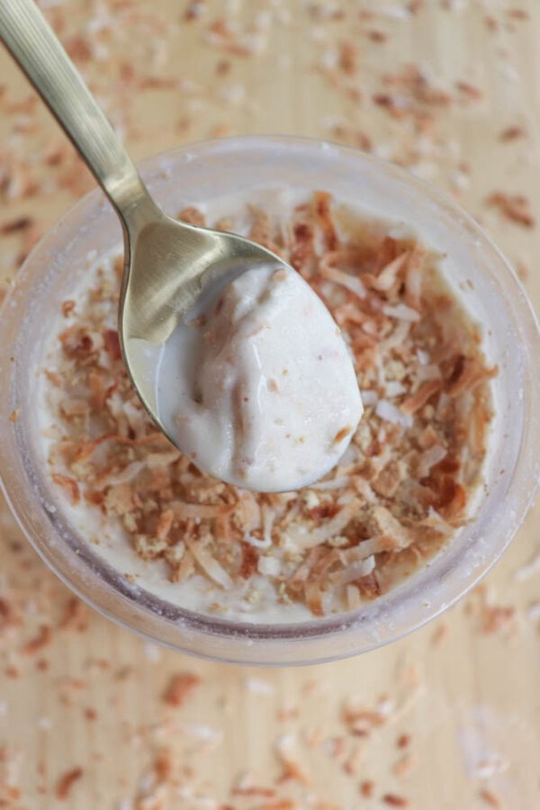 A spoon dips into a bowl of creamy coconut cream pie ice cream topped with toasted coconut flakes. The golden spoon is held above the bowl, which sits on a light wooden surface sprinkled with more coconut flakes.
