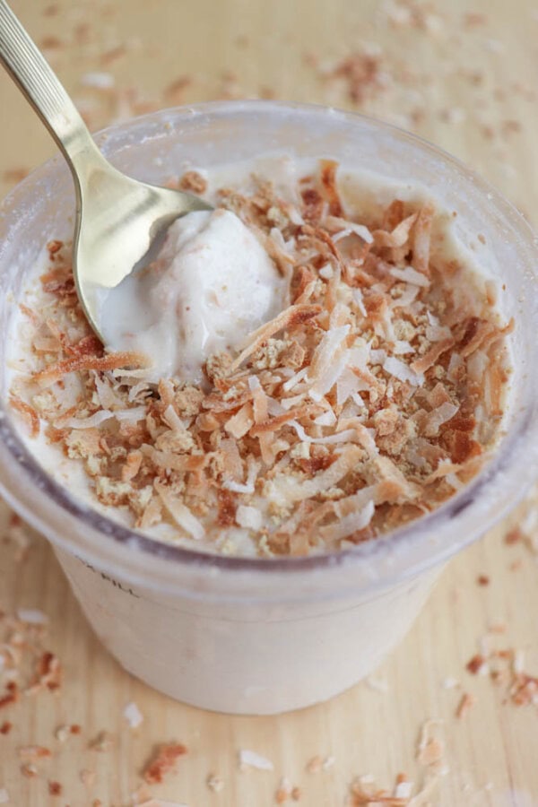 A clear plastic cup filled with creamy coconut cream pie ice cream topped with toasted coconut shavings. A gold spoon rests on top, scooping up a portion. Toasted coconut pieces are scattered around on the wooden surface below.