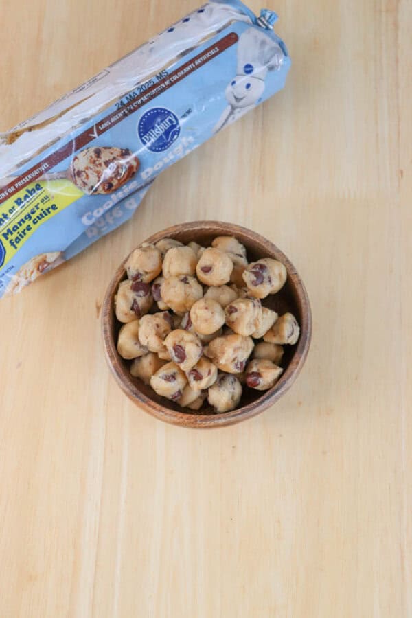 A bowl filled with chocolate chip cookie dough pieces sits on a wooden surface. Behind the bowl, there is an open package of Pillsbury Cookie Dough, displaying the Pillsbury logo and cookie images.