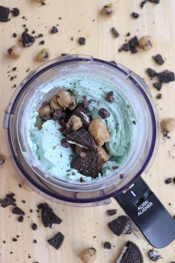 A ninja creami cup filled with light blue ice cream, topped with cookie dough chunks and crushed chocolate cookies. Additional cookie crumbs and dough pieces are scattered on the light wooden surface around it.