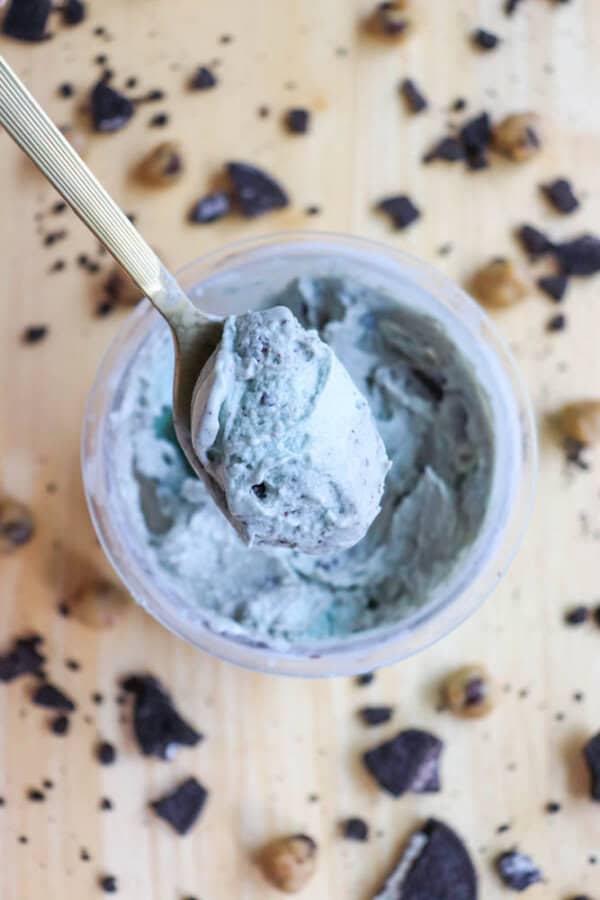 A spoon holding a scoop of cookies and cream ice cream with chocolate chunks, above an open container. Crumbled cookies and cookie dough pieces are scattered on a wooden surface around the container.