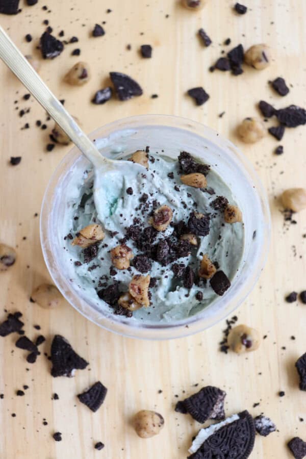 A top view of a bowl containing light blue ice cream mixed with chunks of cookies and small pieces of cookie dough. A gold spoon is in the bowl. Crushed cookies and dough pieces are scattered on the wooden surface around it.