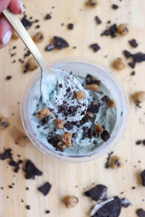A spoonful of mint chocolate chip ice cream with cookie crumbles and chocolate chunks is being lifted from a container. Scattered cookie pieces and chocolate chips are on the wooden surface around the container.