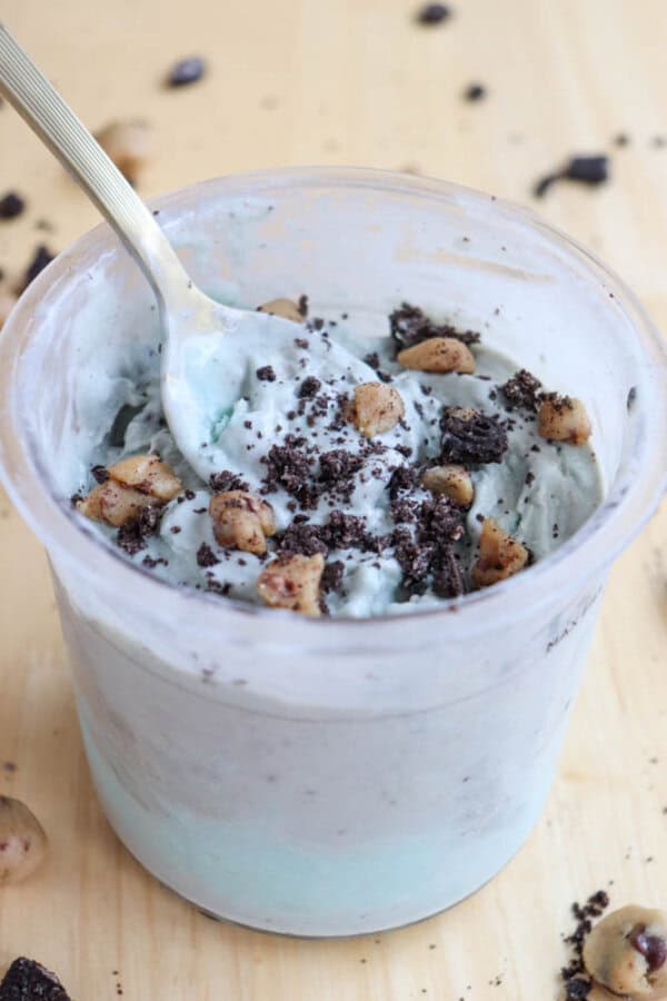 A cup of cookie monster ice cream, topped with crumbled cookies and chocolate chip dough pieces. A spoon is dipped into the creamy dessert, with a wooden surface visible in the background.